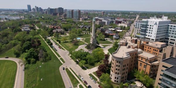 Aerial view of a city park with a central monument, surrounded by buildings, trees, and roads, with a skyline of high-rise buildings in the background.
