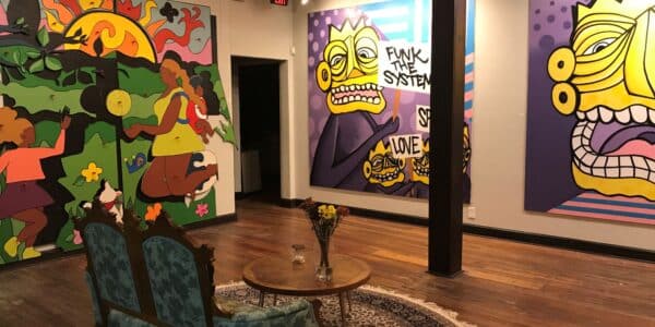 Art gallery with colorful, abstract murals on the walls, wooden floors, a vintage loveseat, and a small table with dried flowers in the center of the room.