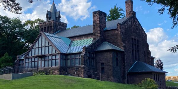 Stone building with steep gabled roofs, a small tower, and large windows, set on a grassy hill with trees and a partly cloudy sky in the background.