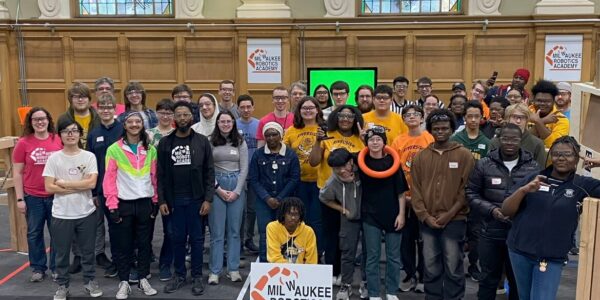A diverse group of students and mentors pose in a gym with “Milwaukee Robotics Academy” signs, robotics equipment, and stained glass windows in the background.