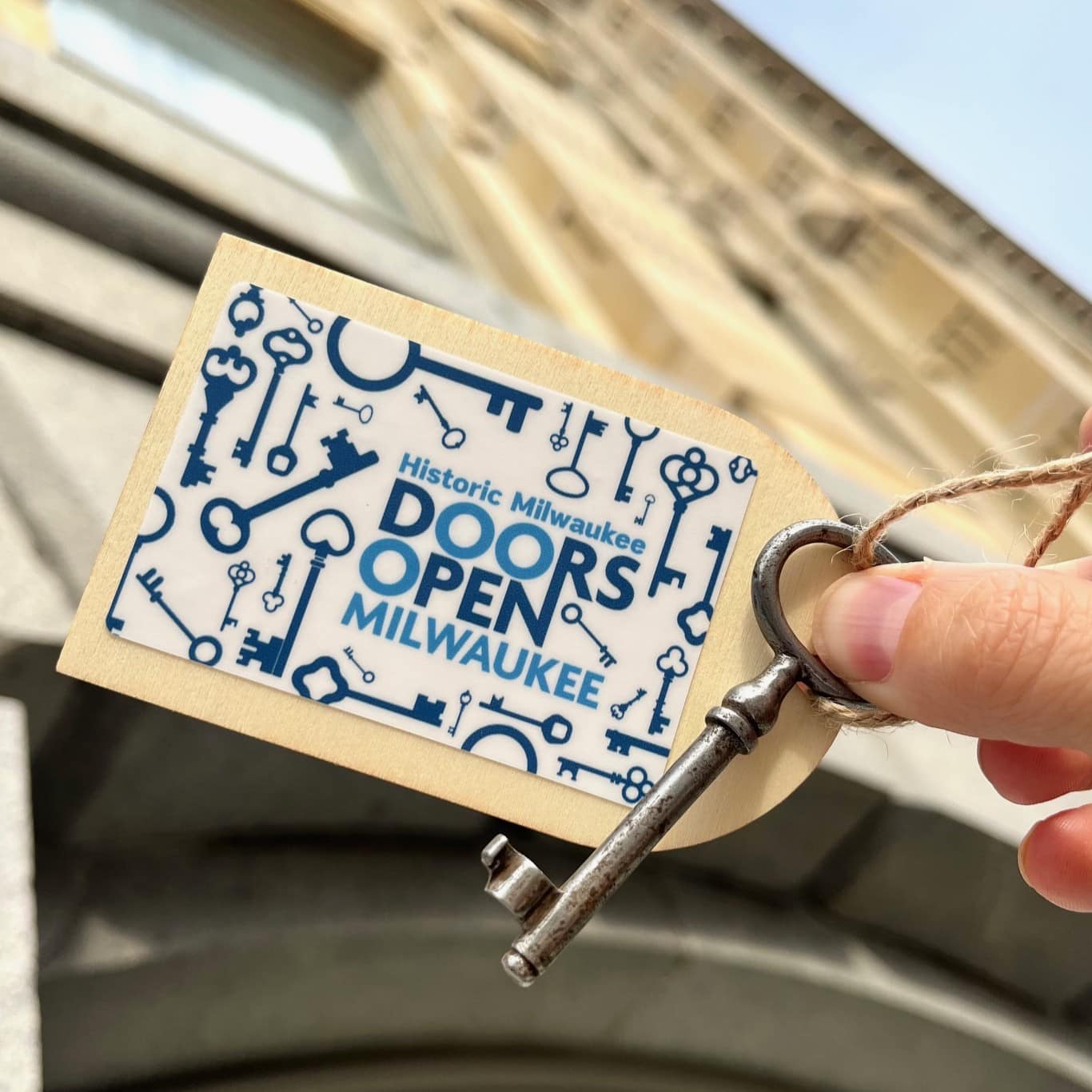 A hand holds an old-fashioned key with a tag that reads "Historic Milwaukee Doors Open Milwaukee" in front of a historic building.