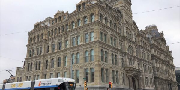 A modern streetcar passes a large, historic stone building with ornate architectural details on a cloudy day at an urban intersection.