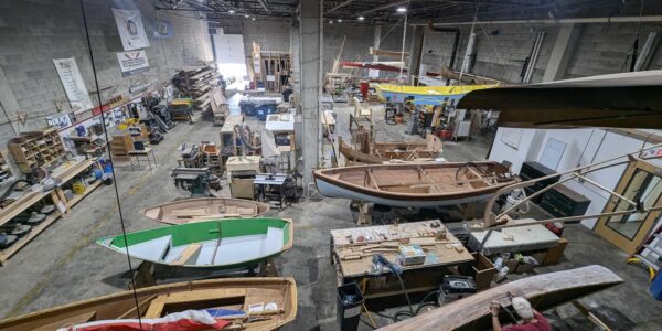 Wide view of a spacious boat workshop with multiple wooden boats in various stages of construction, workbenches, tools, and materials visible throughout the industrial space.