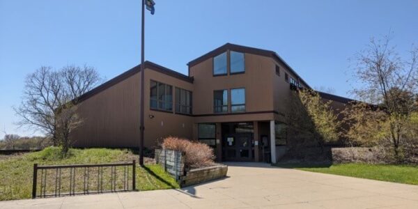A two-story brown building with large windows and a sloped roof stands behind a flagpole under a clear blue sky. Sparse trees and trimmed bushes surround the entrance.