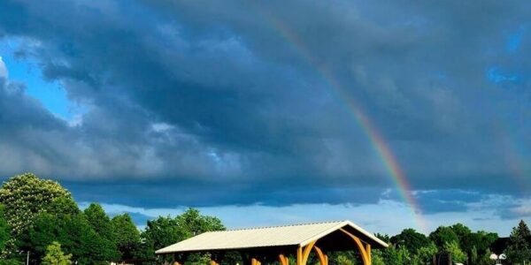 A rainbow arcs over a picnic shelter in a green park, with cloudy skies above and vegetable garden plots in the foreground.