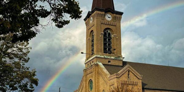 A brick church with a tall clock tower stands on a street corner under a cloudy sky, with a rainbow arching behind it and autumn trees nearby.