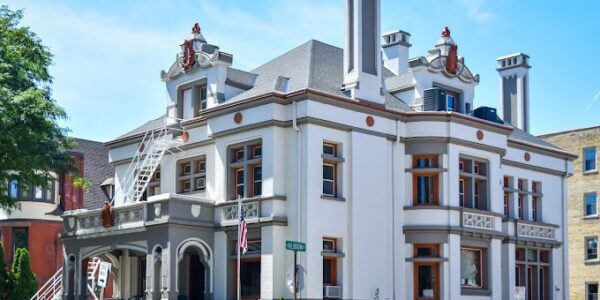 A large, historic two-story building with ornate architectural details, multiple chimneys, and a prominent front porch sits at a street corner on a sunny day.