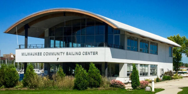 A modern two-story building with large windows labeled “Milwaukee Community Sailing Center,” surrounded by grass, shrubs, and trees under a clear blue sky.