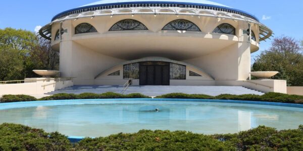 A round, modernist building with large arched windows and a dome roof stands behind a circular reflecting pool and landscaped bushes under a blue sky.