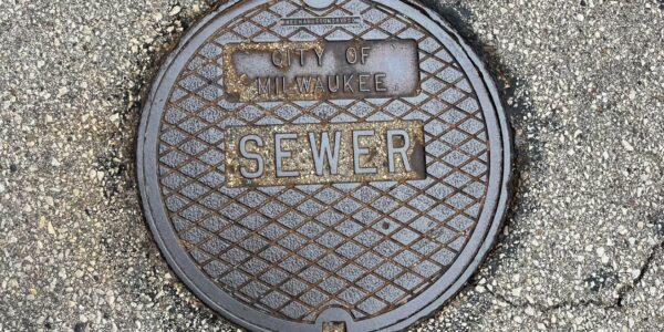 A round metal sewer cover labeled “City of Milwaukee SEWER” set in cracked, textured pavement.