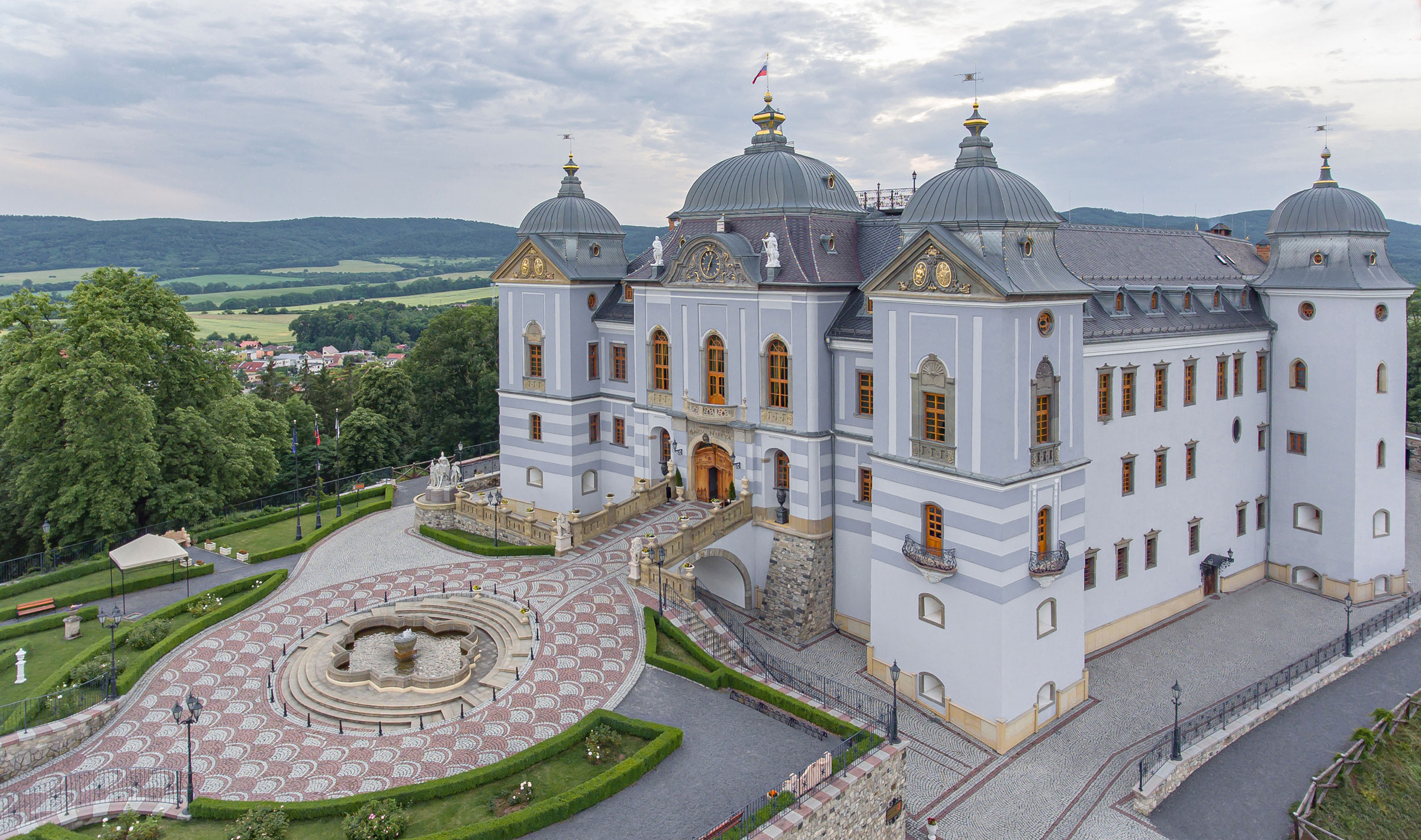 Large, ornate palace with gray domes, central fountain, and landscaped grounds, set against a backdrop of green hills and cloudy sky.