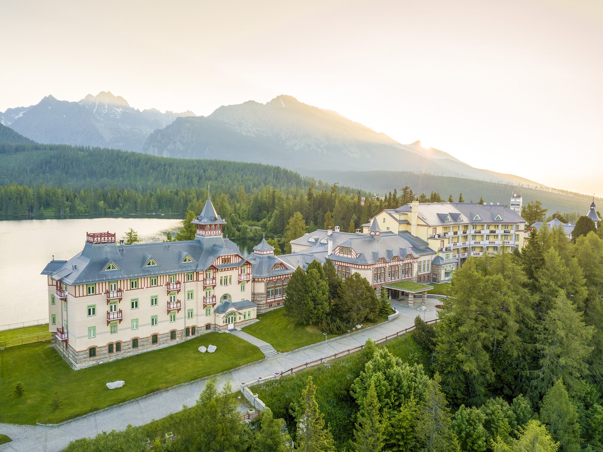 Large lakeside hotel complex surrounded by trees, with mountains in the background and the sun rising or setting behind them.