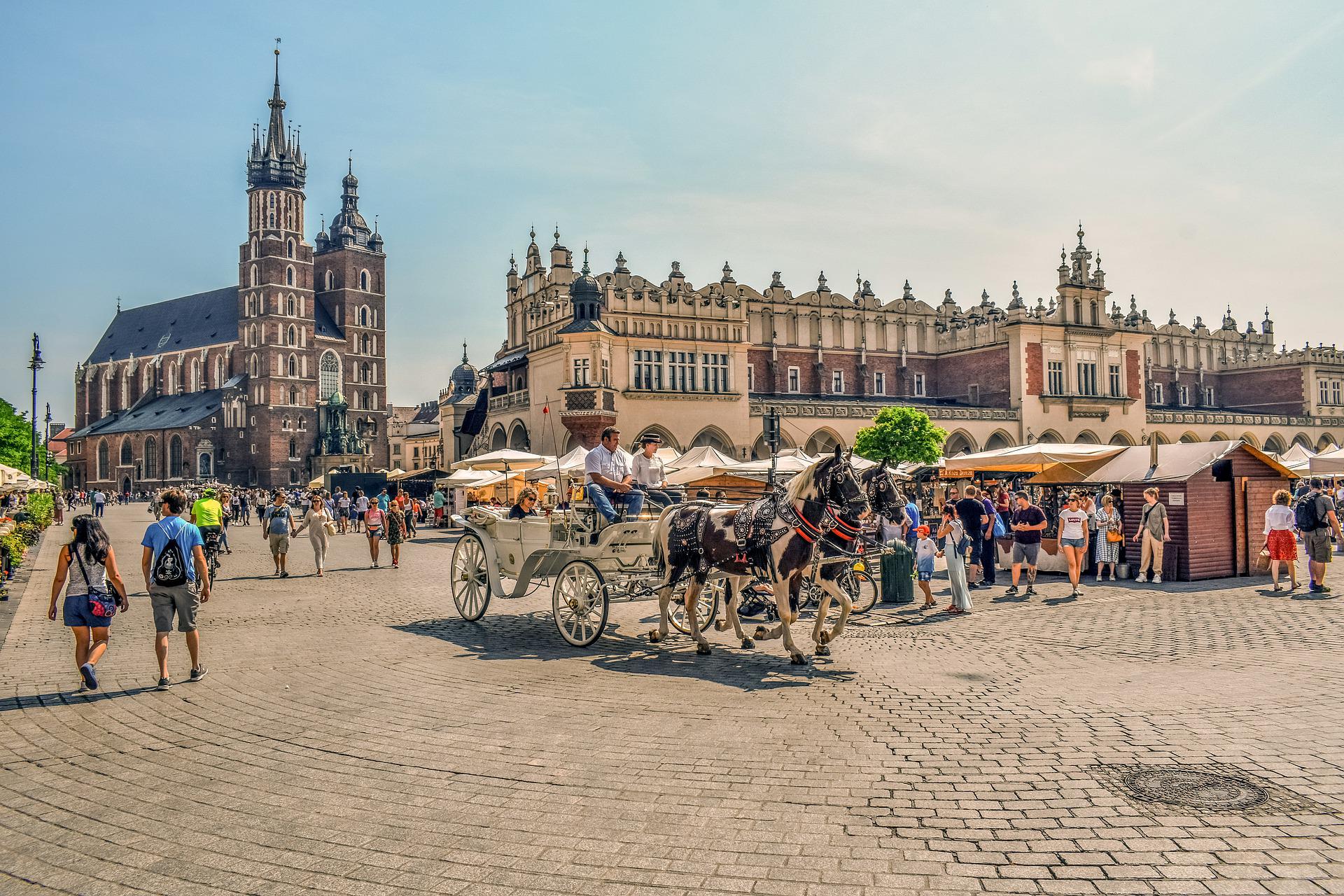 A horse-drawn carriage passes through a busy city square with historic buildings, street vendors, and pedestrians on a sunny day.