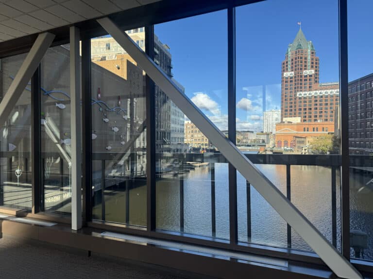 View of a city river and surrounding buildings through large glass windows with metal beams, taken from inside a pedestrian walkway.