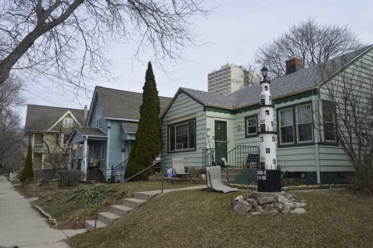 A row of houses lines a residential street; one yard features a large white and black model lighthouse surrounded by rocks.