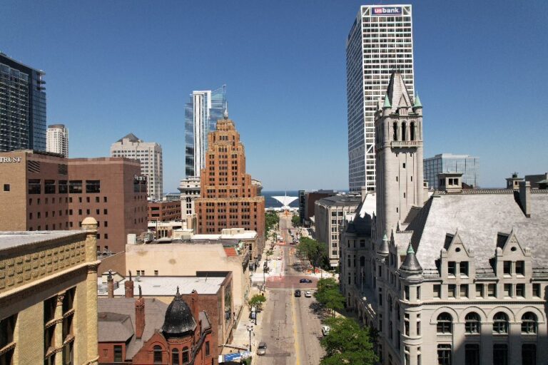 A cityscape view of downtown Milwaukee, Wisconsin, showing tall buildings, historic architecture, and Lake Michigan in the background under a clear blue sky.