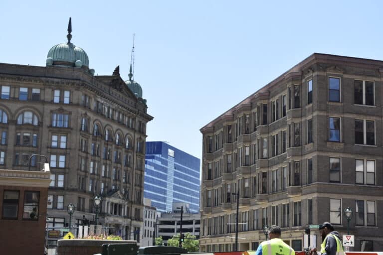 Two construction workers stand in front of historic and modern buildings under a clear blue sky in an urban setting.