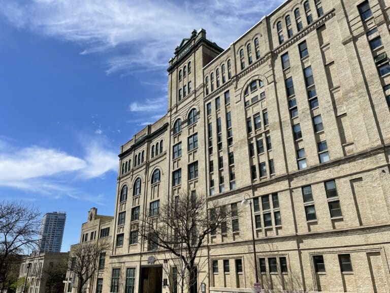 A large, multi-story historic building with light brick exterior, arched windows, and a central tower stands under a partly cloudy blue sky. Leafless trees line the sidewalk in front.