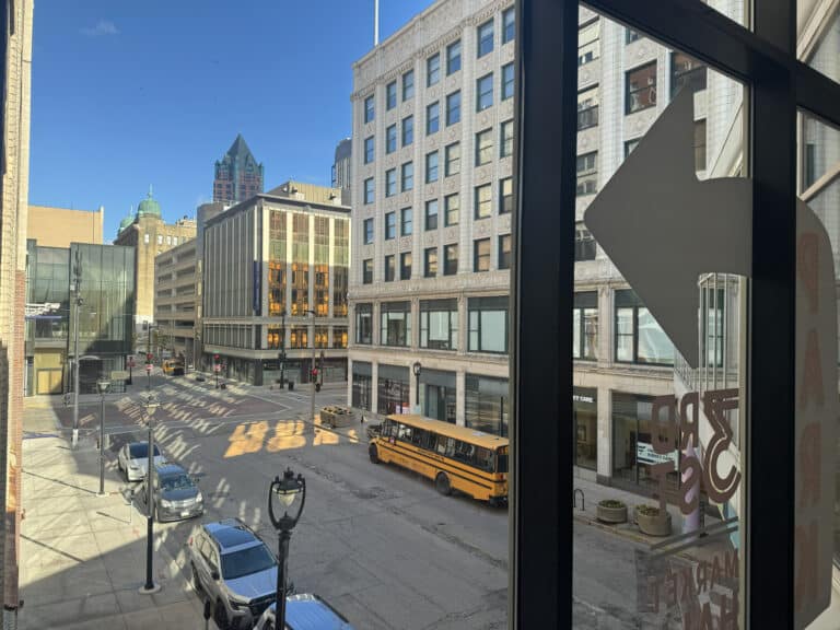 A view through large windows shows a downtown street with a yellow school bus, light traffic, and several multi-story buildings on a sunny day.