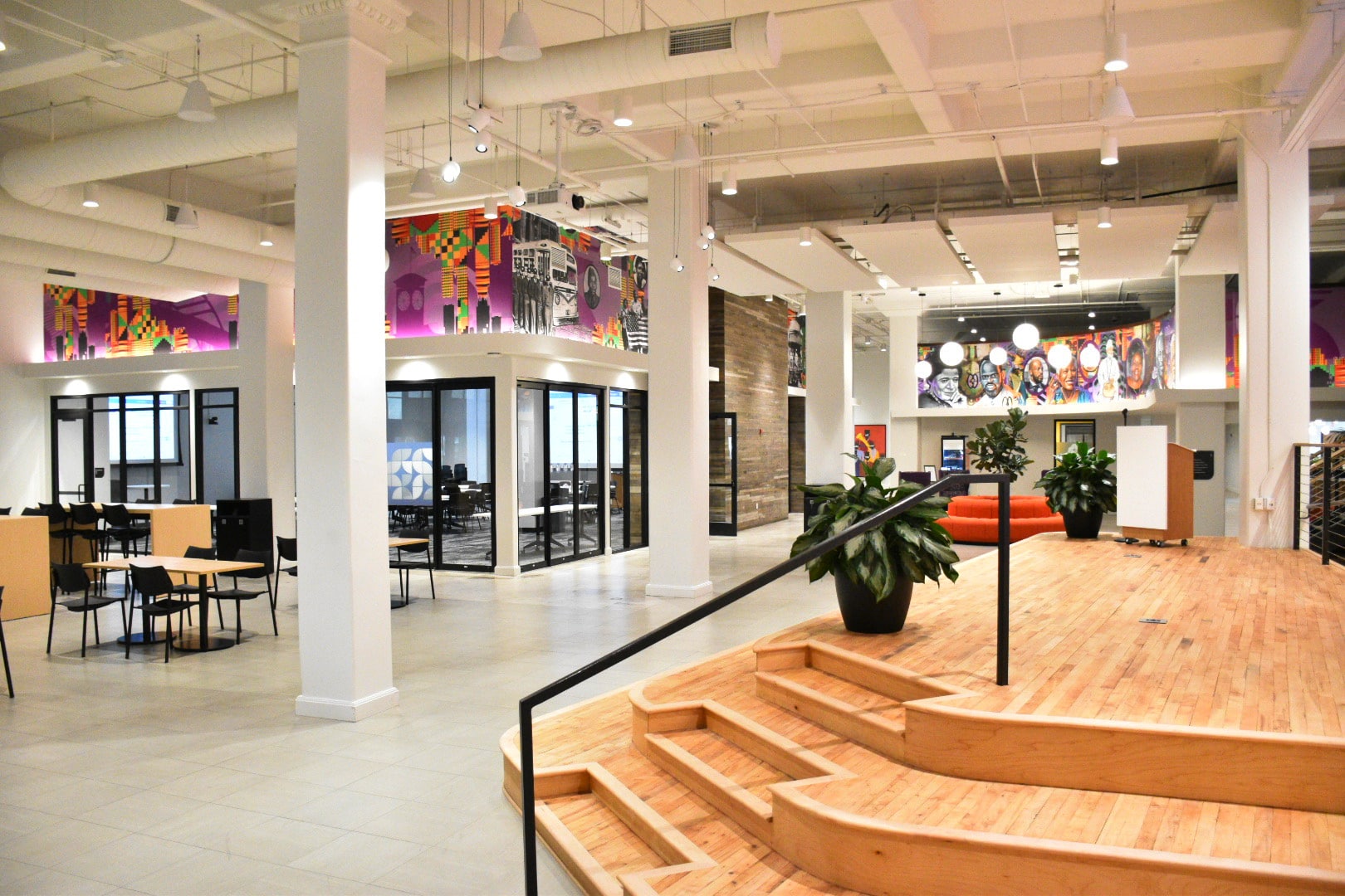 Modern open-plan office space with wooden steps, glass offices, colorful murals on the walls, tables, chairs, and potted plants.