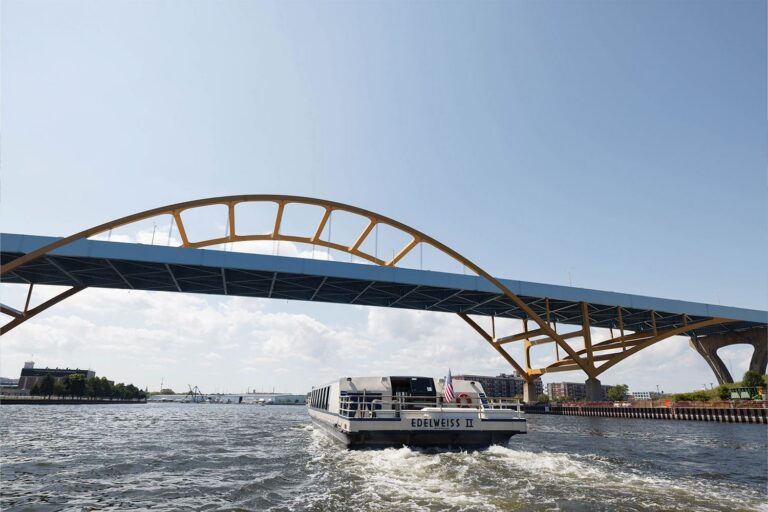 A white boat labeled "EDELWEISS II" travels under a large yellow-arched bridge on a sunny day with scattered clouds.