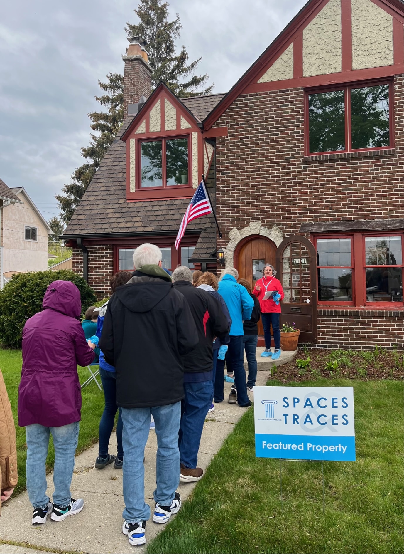 A group of people stand in line outside a brick house with an American flag, near a sign reading "Spaces Traces Featured Property.
