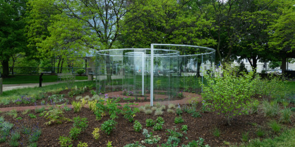 A circular outdoor glass maze stands in a landscaped garden surrounded by trees and shrubs on a cloudy day.