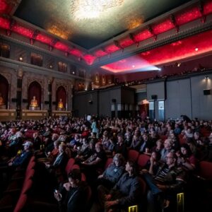 A large audience sits in a dimly lit theater with ornate décor and red ceiling lights, focusing on an unseen stage or screen.