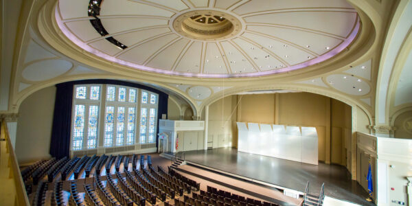 Large, empty auditorium with arched windows, rows of brown seats, and a stage under a domed ceiling with decorative molding and recessed lighting.