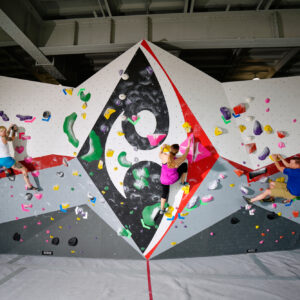 Three people are climbing on a large indoor bouldering wall with colorful handholds and geometric patterns under an exposed metal ceiling.