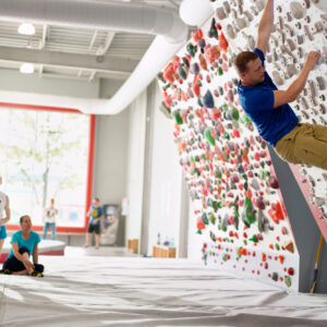 A man climbs an indoor bouldering wall while four people watch him from the padded floor in a brightly lit gym.