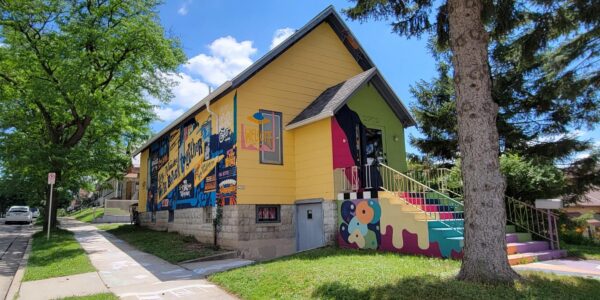 A yellow house with colorful murals and designs on the walls and stairs, surrounded by trees, under a blue sky with clouds.
