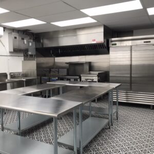 Commercial kitchen with stainless steel counters, industrial refrigerators, stove and ventilation hood, and patterned tile floor.