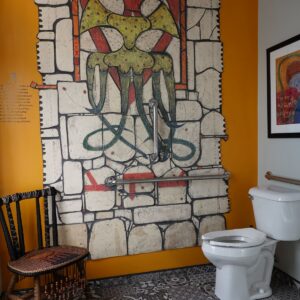A bathroom with patterned tile floor, orange walls, a toilet, a woven chair, and a mural resembling medieval stonework with an abstract, multi-colored creature.
