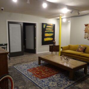 A living room with a yellow couch, two abstract paintings, a wooden coffee table, rugs, and various decorative items on a concrete floor.