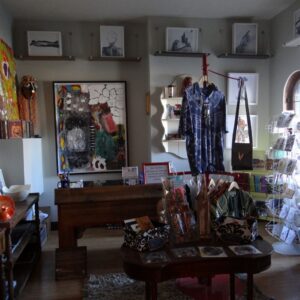 Art gallery with eclectic paintings, sculptures, photography, and crafts displayed on walls, tables, shelves, and racks under natural and artificial light.