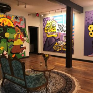 Colorful art gallery with wooden floors, vivid murals on walls, vintage green loveseat, small round table, and dried flowers in a vase at the center.