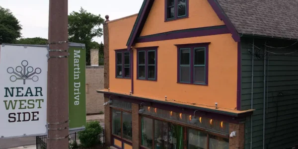 Two people sit on the front steps of a two-story orange and green house with large windows. A street sign reads “Near West Side Martin Drive.”.