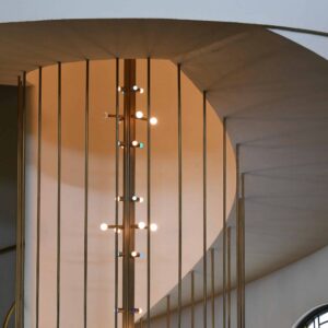 Curved staircase with vertical metal balusters and a modern vertical light fixture, viewed from below, with a round window on the right.