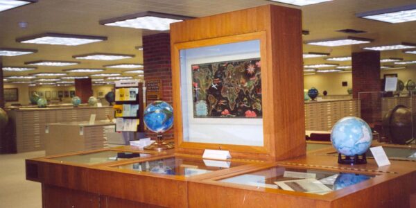 A wooden display case holds a framed map or artwork, with two globes and documents underneath, in a well-lit library or archive room.