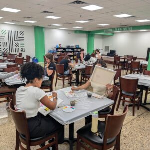 People are seated at tables in a large room, engaged in painting activities with art supplies on each table. Shelves with materials are visible in the background.