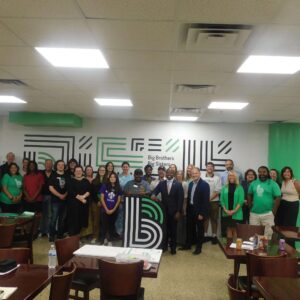 Group of people standing and posing for a photo inside a room with Big Brothers Big Sisters branding on the wall behind them.