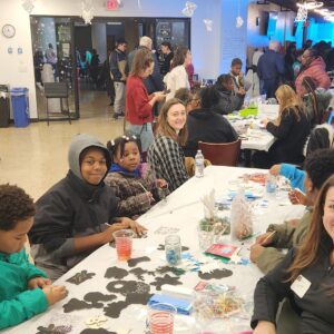 People of various ages sit at tables doing crafts in a busy, well-lit community center. Some smile for the camera, while others focus on their activities. Decorations hang from the ceiling.