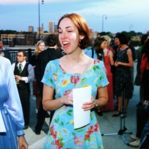 A woman in a floral dress holds a folded paper while talking with others at an outdoor event; several people are gathered in the background.