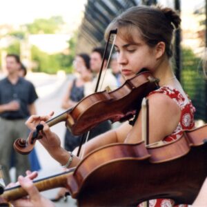 Two women play violins outdoors, reading sheet music, with a group of people and a fence in the blurred background.