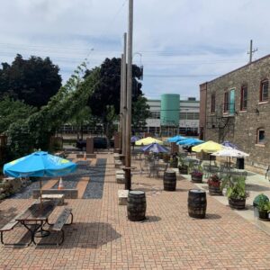 Outdoor patio with brick flooring, picnic tables, colorful umbrellas, wooden barrels, and potted plants near a brick building on a sunny day.