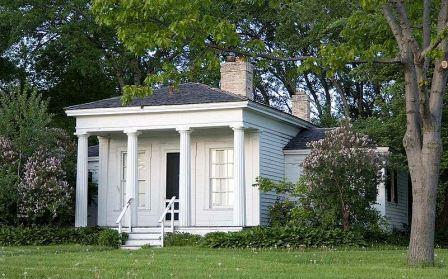 Small white house with four columns, black roof, and two chimneys, surrounded by green trees and shrubs on a grassy lawn.