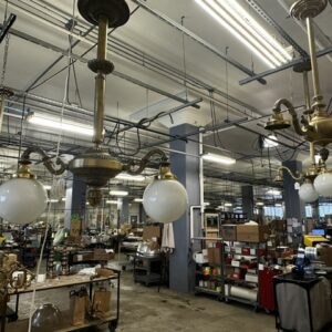 A workshop with hanging brass chandeliers, fluorescent lights, shelves with boxes, and various tools and equipment scattered throughout the space.