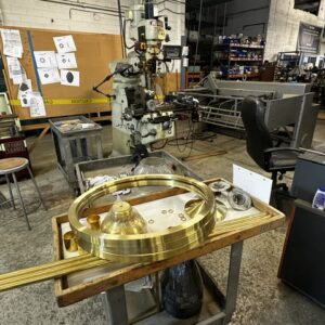 A metal workshop with machining equipment, blueprints on the wall, and brass parts on a cart in the foreground.