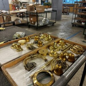 Tray with assorted brass light fixture parts on a worktable in an industrial workshop, with shelves and boxes visible in the background.